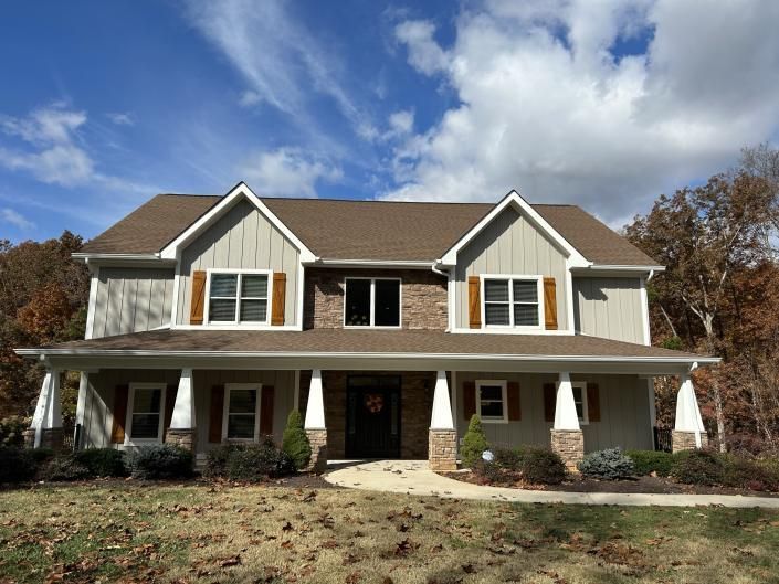 Two-story suburban house with beige siding, brown roof, white columns, and a front porch under a blue sky