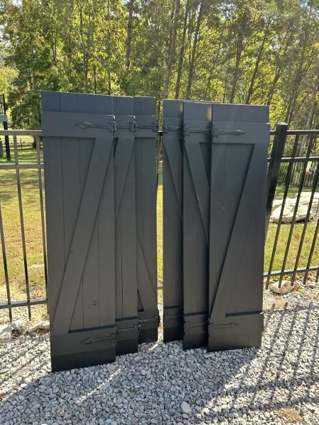 Two dark metal gate panels leaning against a black fence on gravel, with trees in the background