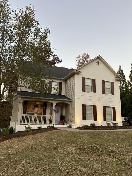 Two-story cream house with a front porch, lit windows, and autumn trees at dusk