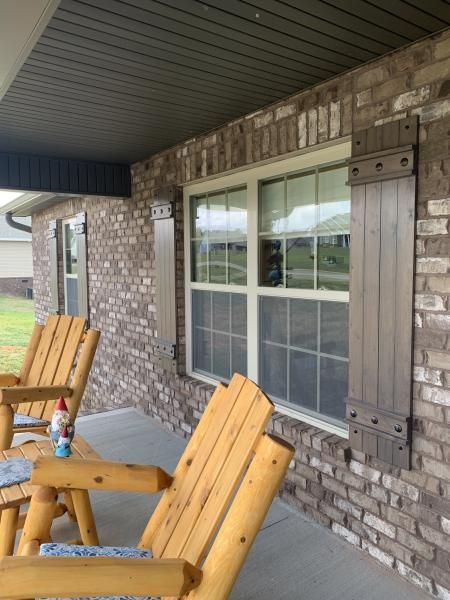 Covered brick porch with large window, brown shutters, and wooden chairs.