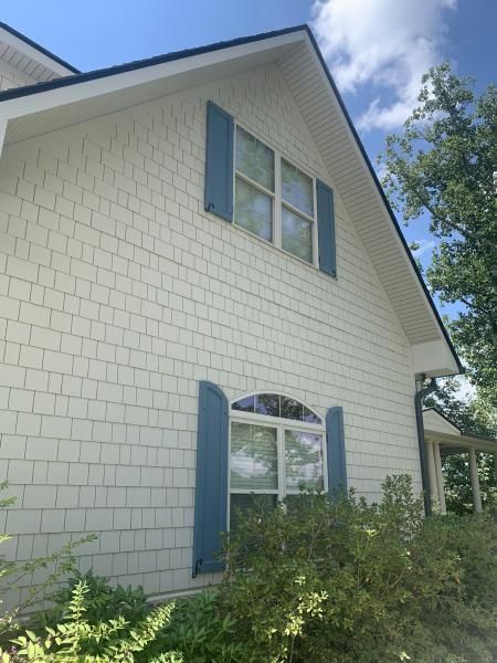 White house exterior with blue shutters and arched window, partly framed by shrubs and trees