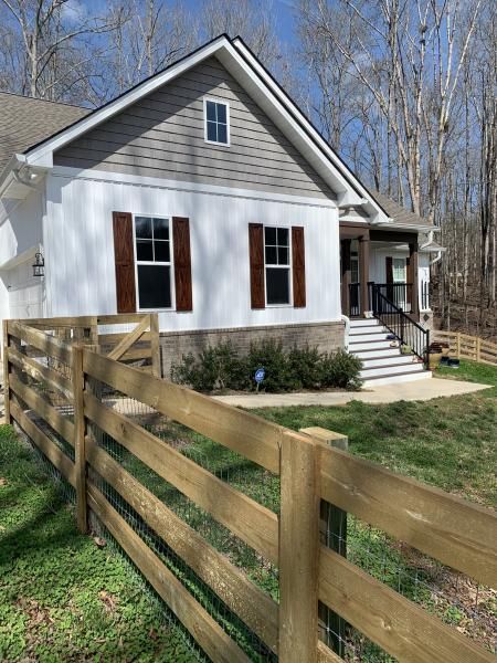 White house with red shutters and a front porch, viewed from behind a wooden fence on a sunny day