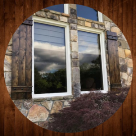 Stone house wall with two windows reflecting clouds and trees, framed by wood trim and shrubs below