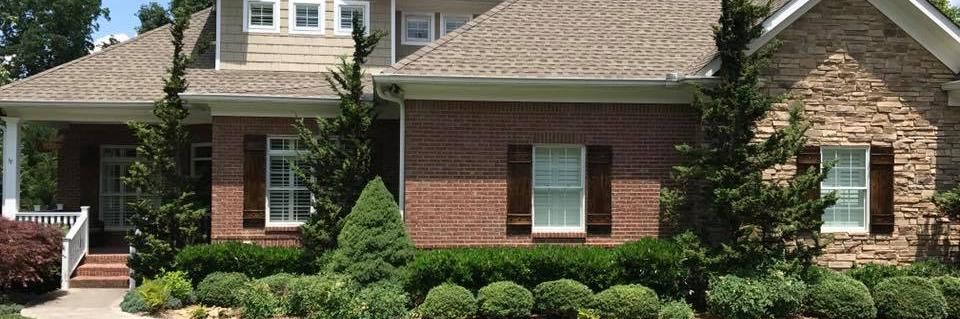 Brick suburban house with tan roof, white windows, and manicured shrubs in front yard
