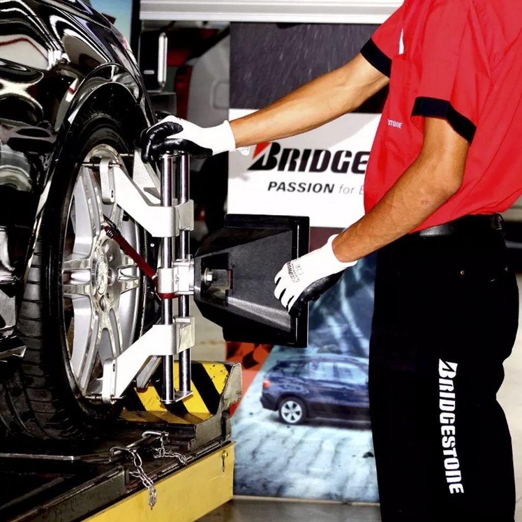 Un mecánico con camisa roja y guantes ajusta la alineación de las ruedas de un coche. El escenario es un taller de neumáticos Bridgestone.