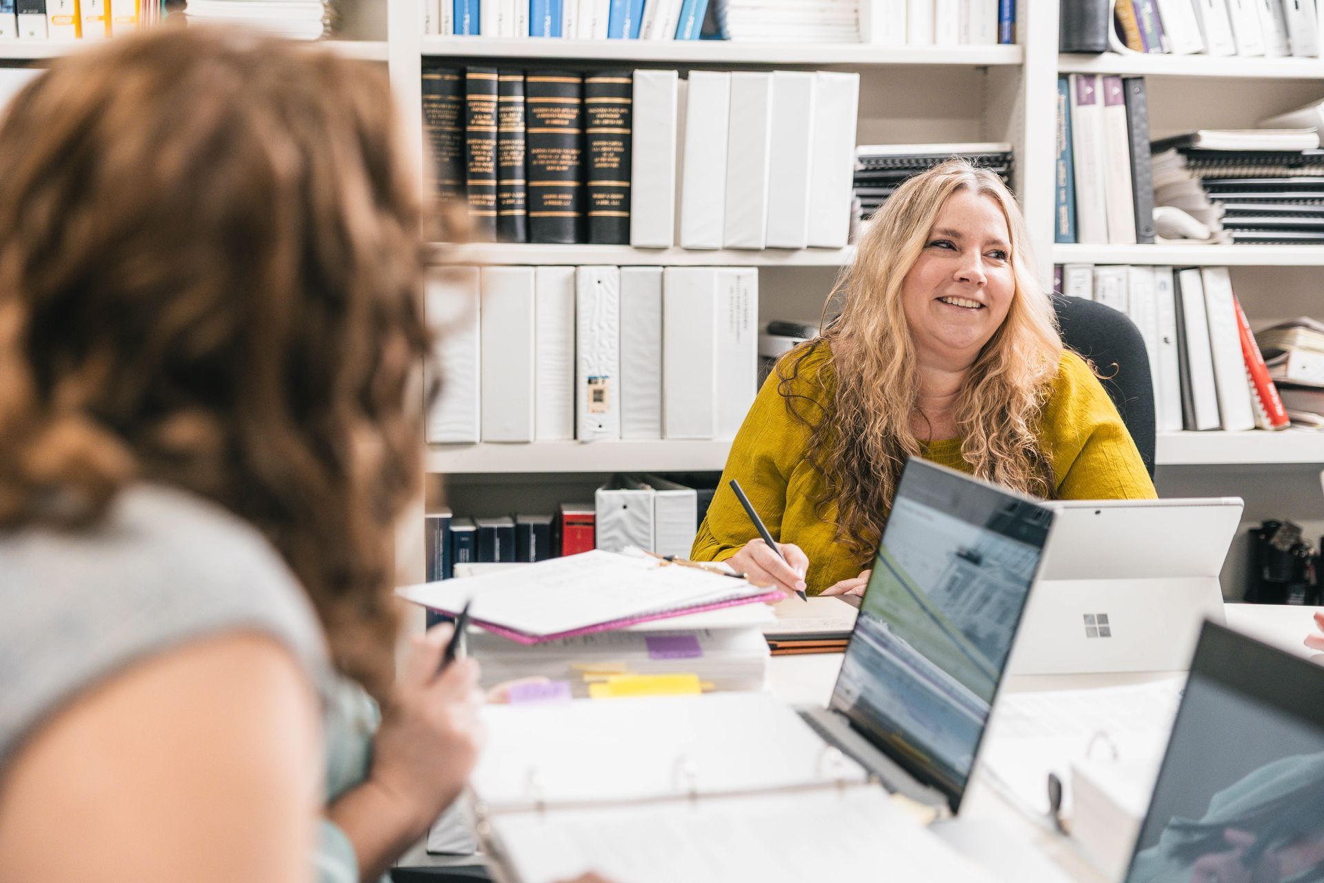 Two women at a desk in an office, one writing and smiling, the other facing away. Shelves with books and binders.