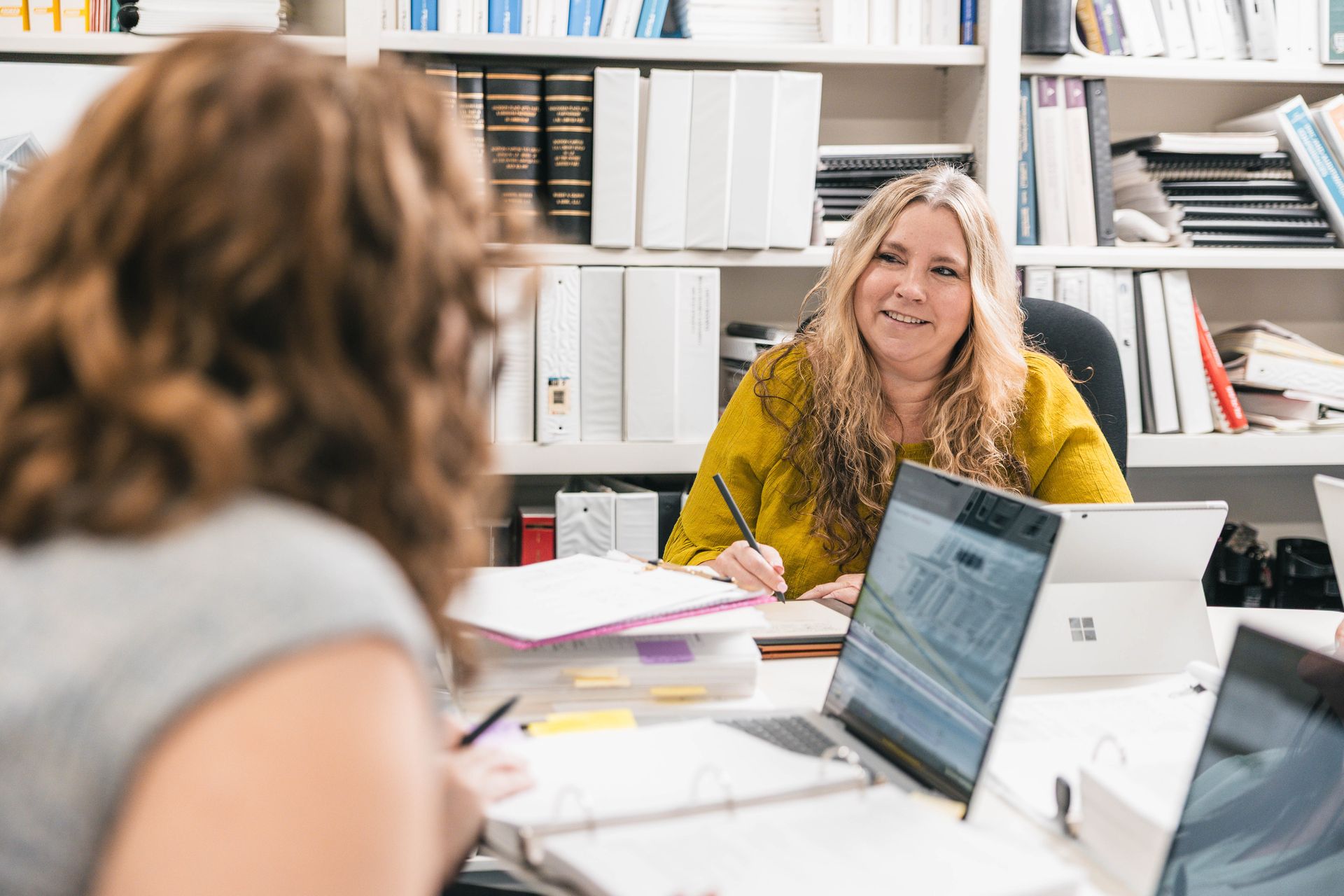 Two people reviewing paperwork at a desk. One points with a pen, other uses a laptop.