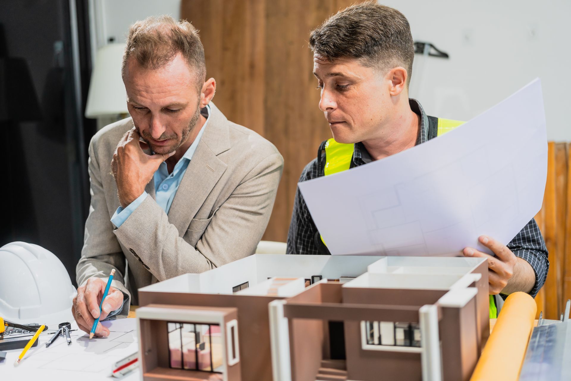 Two men reviewing blueprints and model of a building. One wears a suit, the other a safety vest.