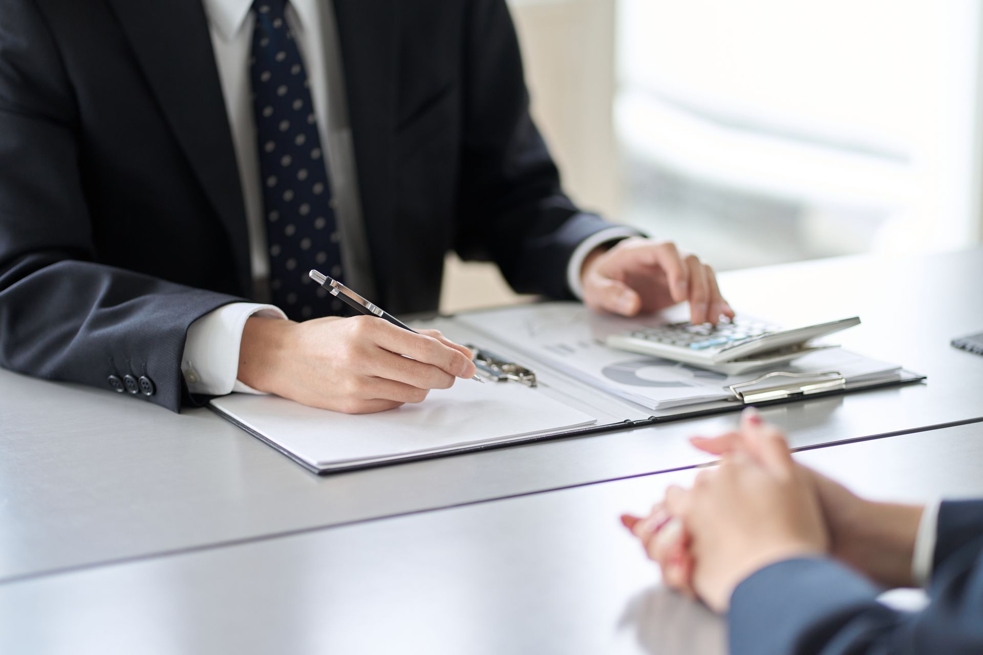 Man in suit writing on document, using calculator; hands clasped in foreground.