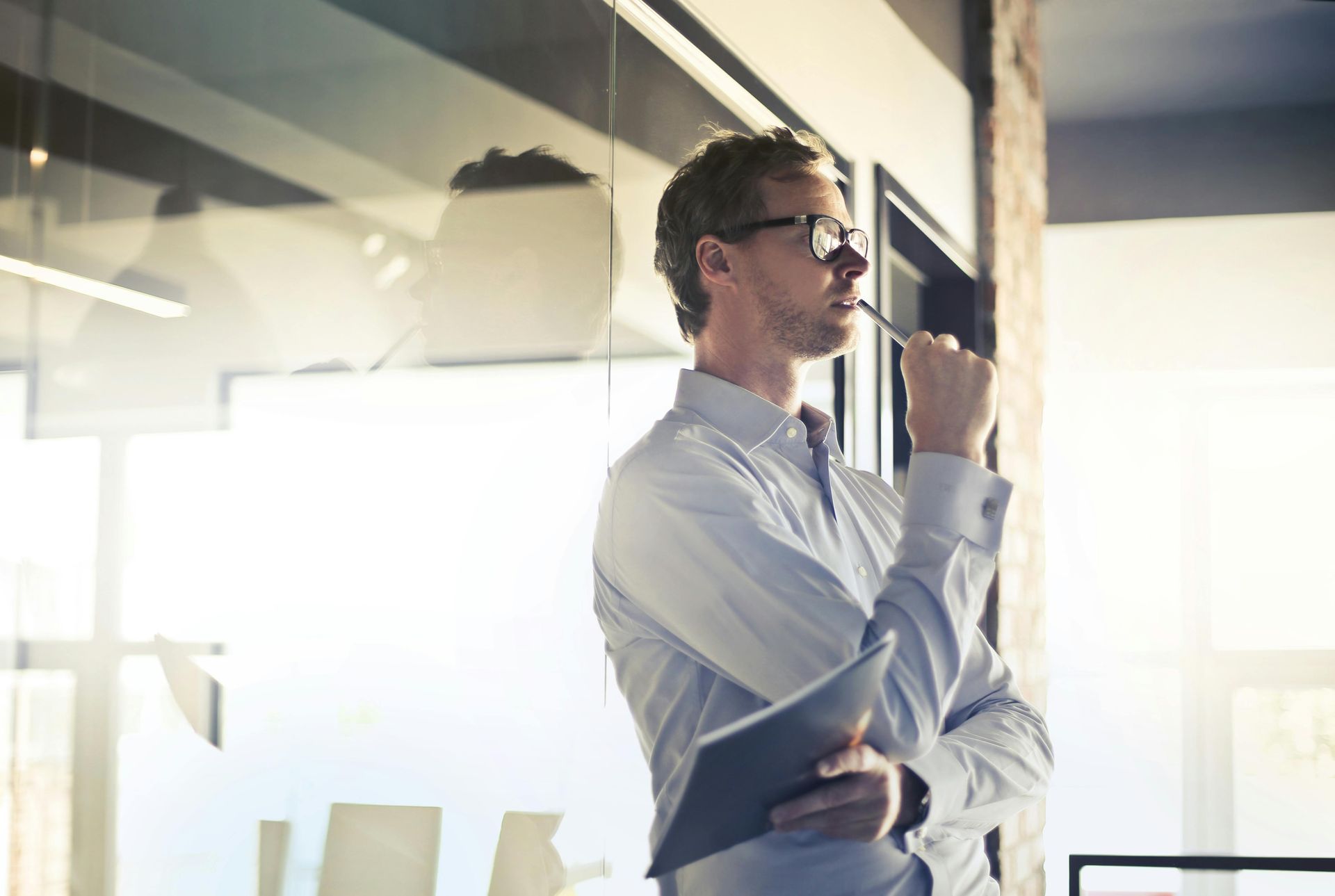 Man in glasses holding a tablet, thinking, leaning against glass wall in office.