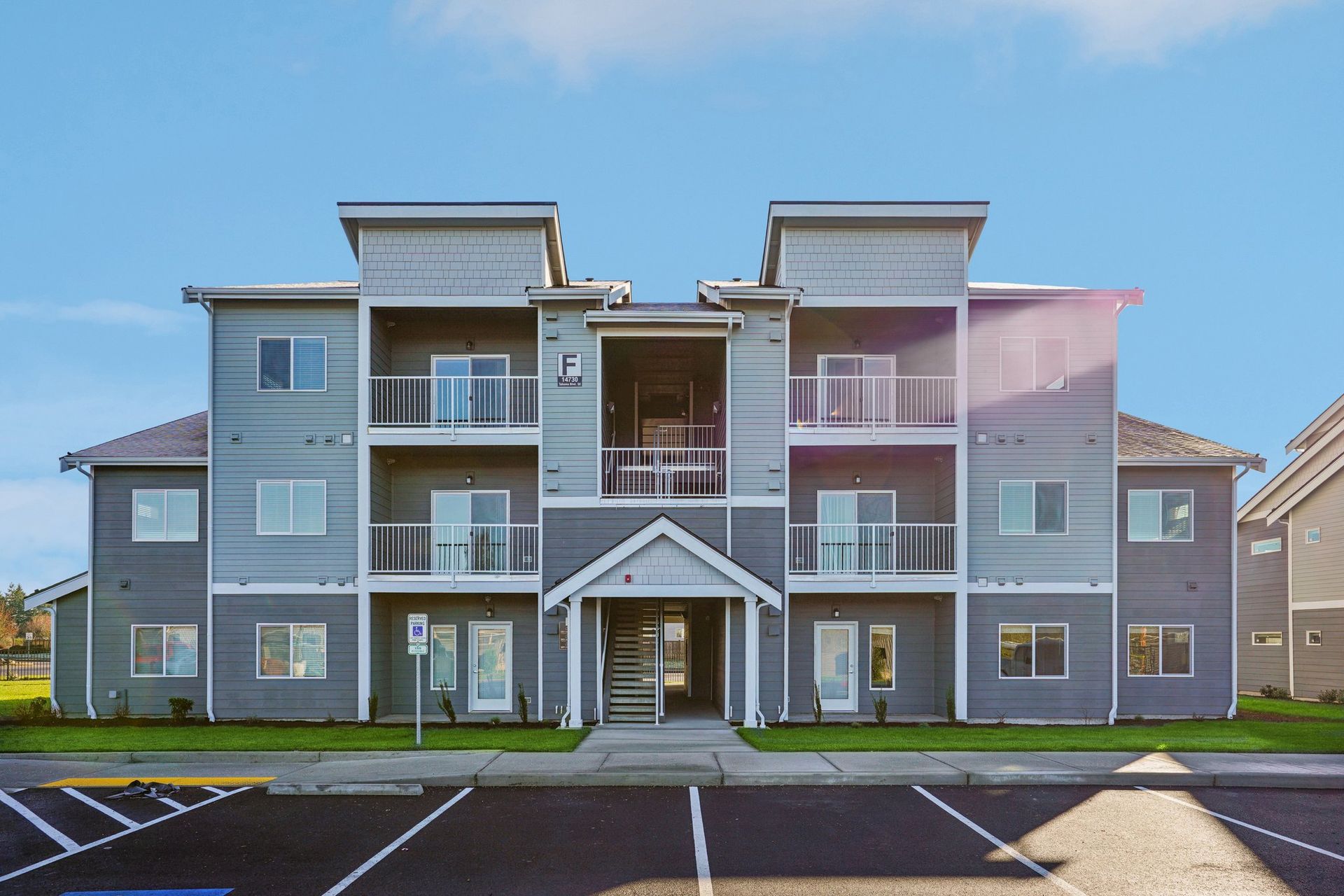 Three-story gray apartment building with balconies and central entrance under a blue sky