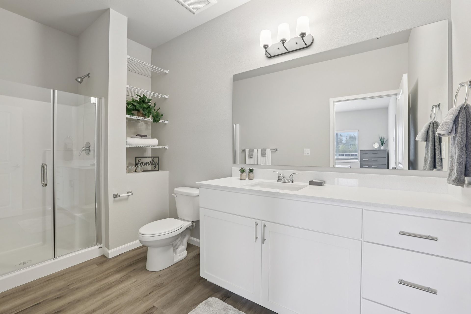Bright modern bathroom with white vanity, large mirror, glass shower, and wood-look flooring.