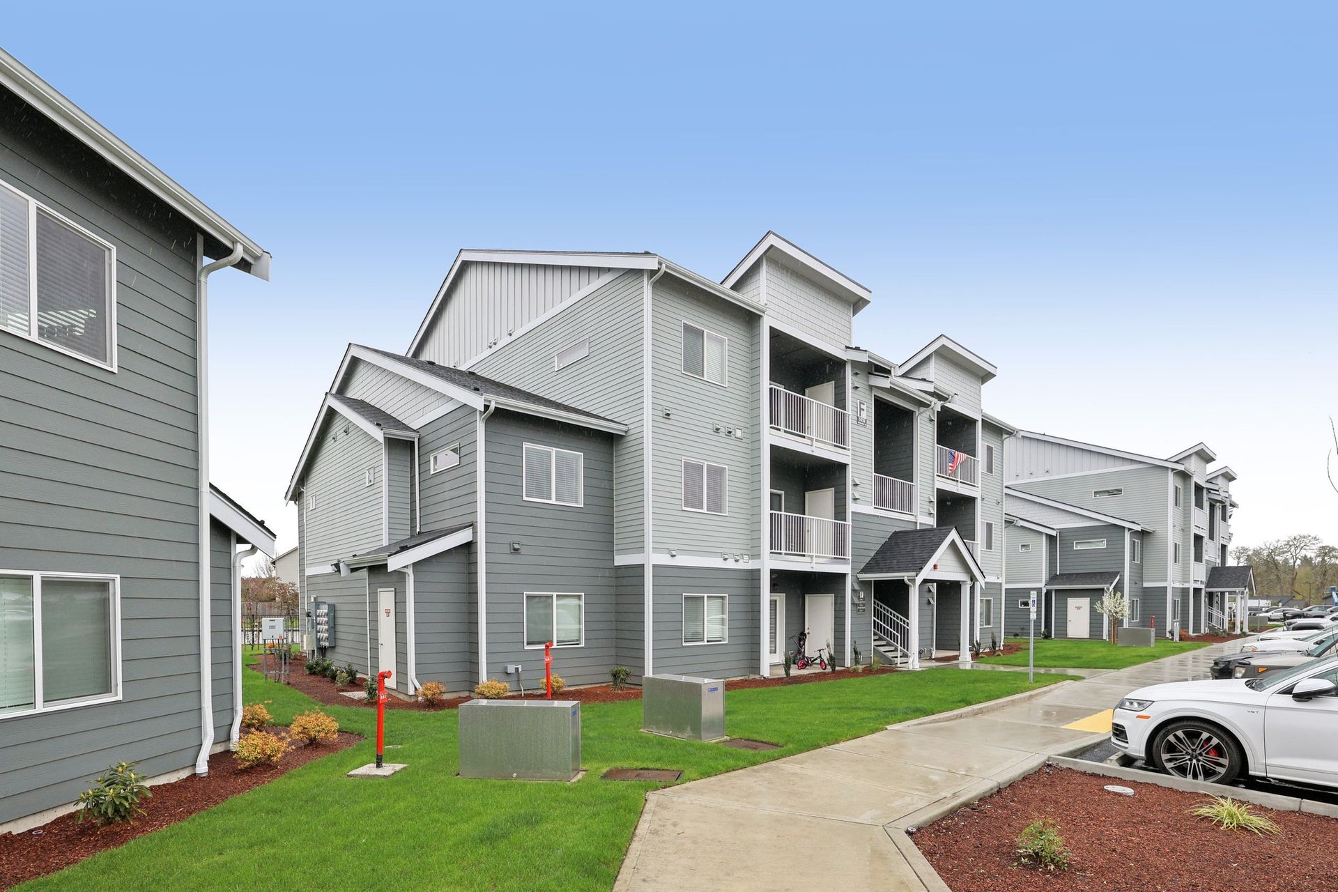 Modern gray apartment buildings with a curved sidewalk, green lawn, and parked cars under a blue sky