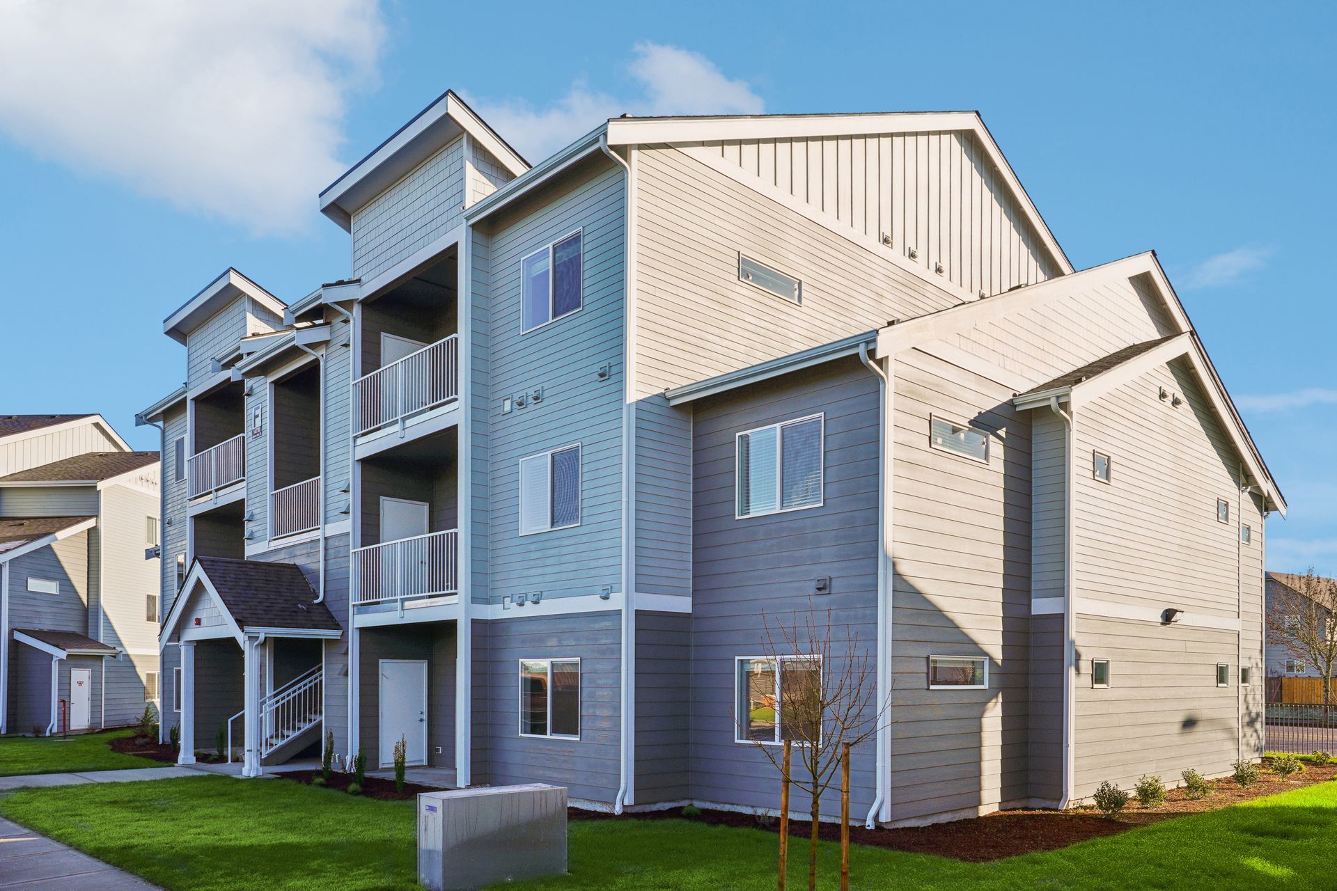 Apartment buildings with gray and beige siding, balconies, and a grassy yard under a clear sky