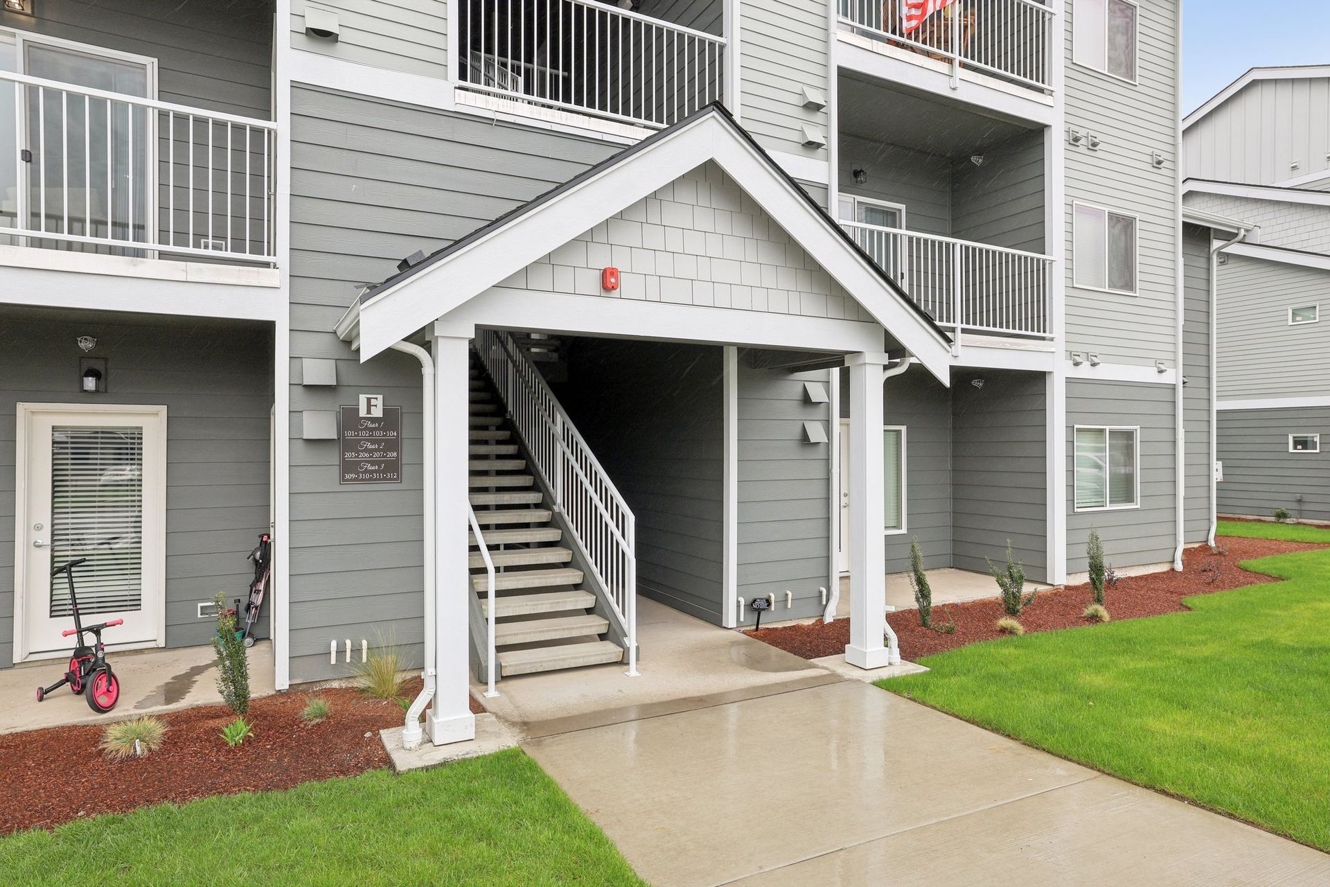 Apartment entrance with stairs and covered walkway, gray siding, concrete path, and green lawn