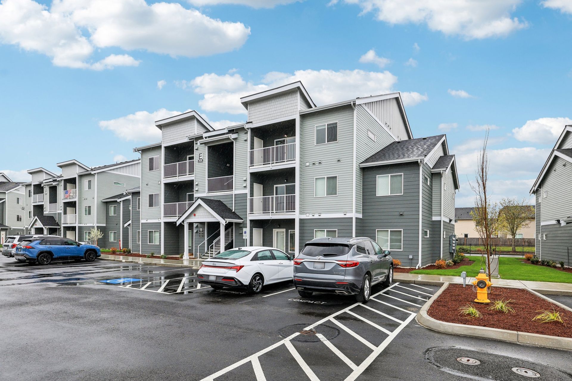 Apartment complex with gray buildings, parked cars, and wet pavement under a cloudy sky