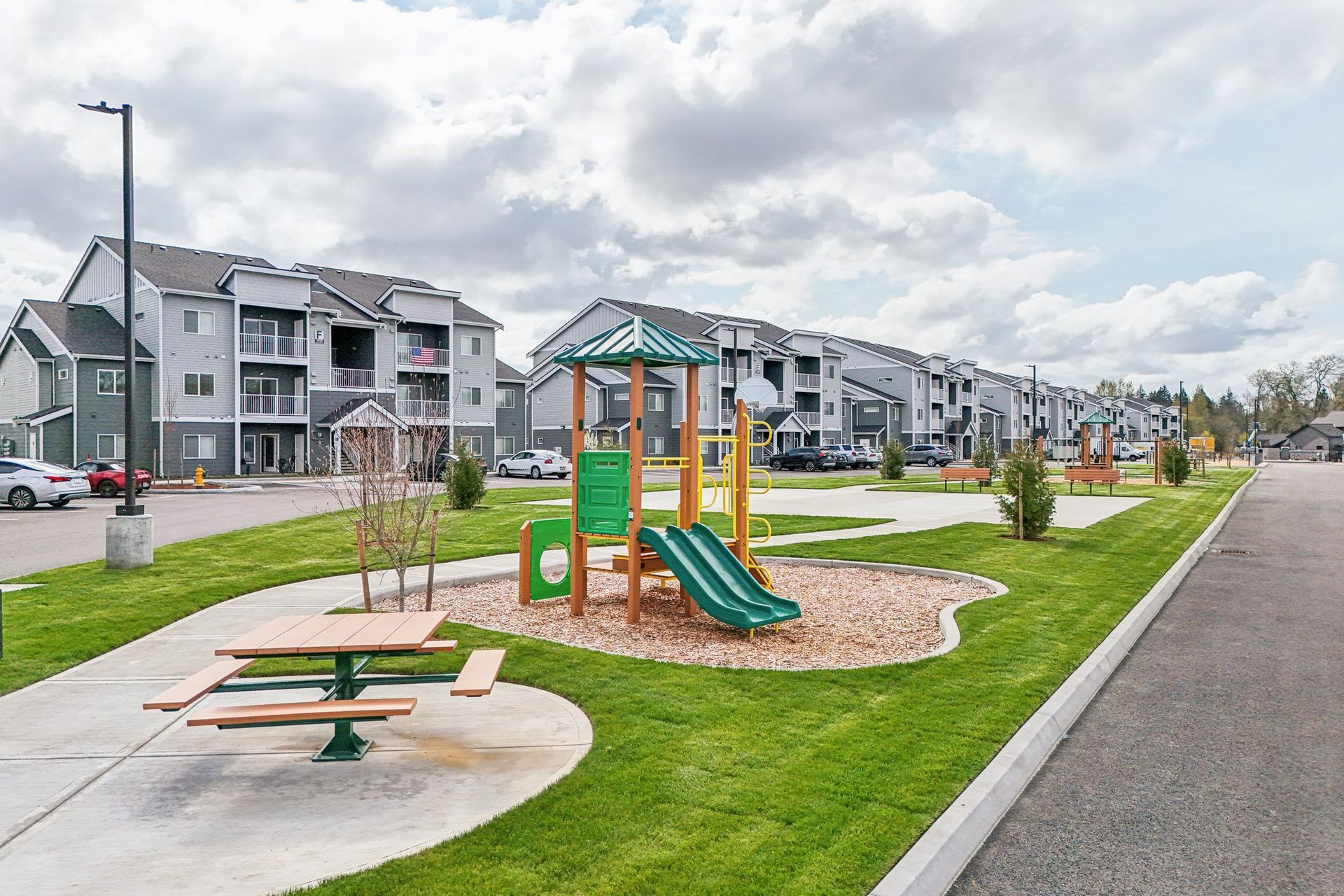 Modern apartment complex with a small playground, picnic table, and paved street under cloudy skies