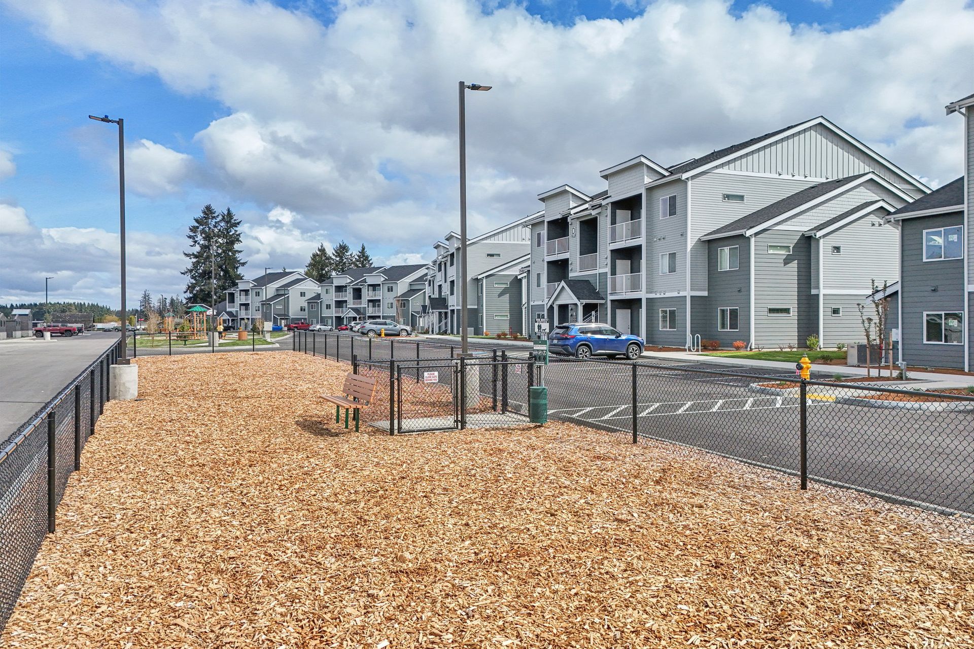 Mulched landscape strip beside apartment buildings and parking lot under a partly cloudy sky