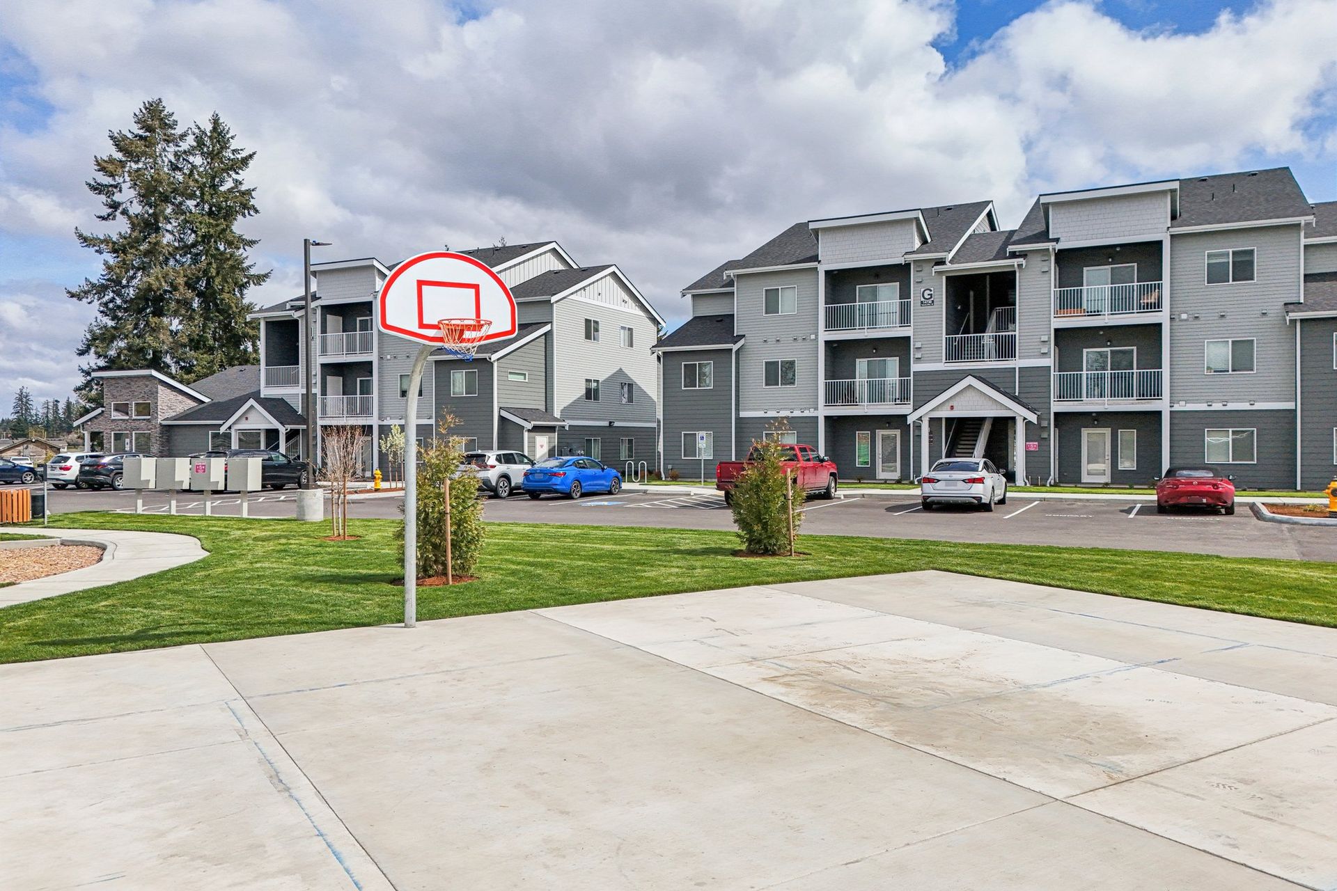 Apartment courtyard with a basketball hoop and concrete court, with parked cars and townhomes in the background