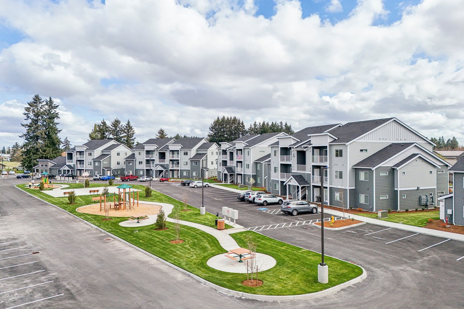 Modern gray apartment complex with landscaped courtyard, road, parked cars, and cloudy sky