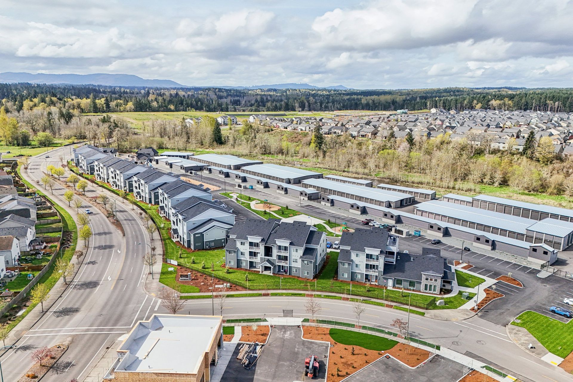 Aerial view of a modern apartment complex along a curved road, with trees and cloudy sky nearby