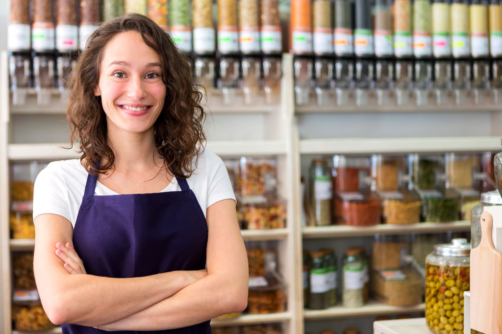 A woman in an apron is standing in a grocery store with her arms crossed.