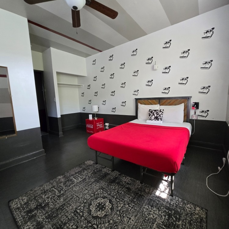 Bedroom with red bedspread, black rug, Coca-Cola cooler, and wall art against a white background.