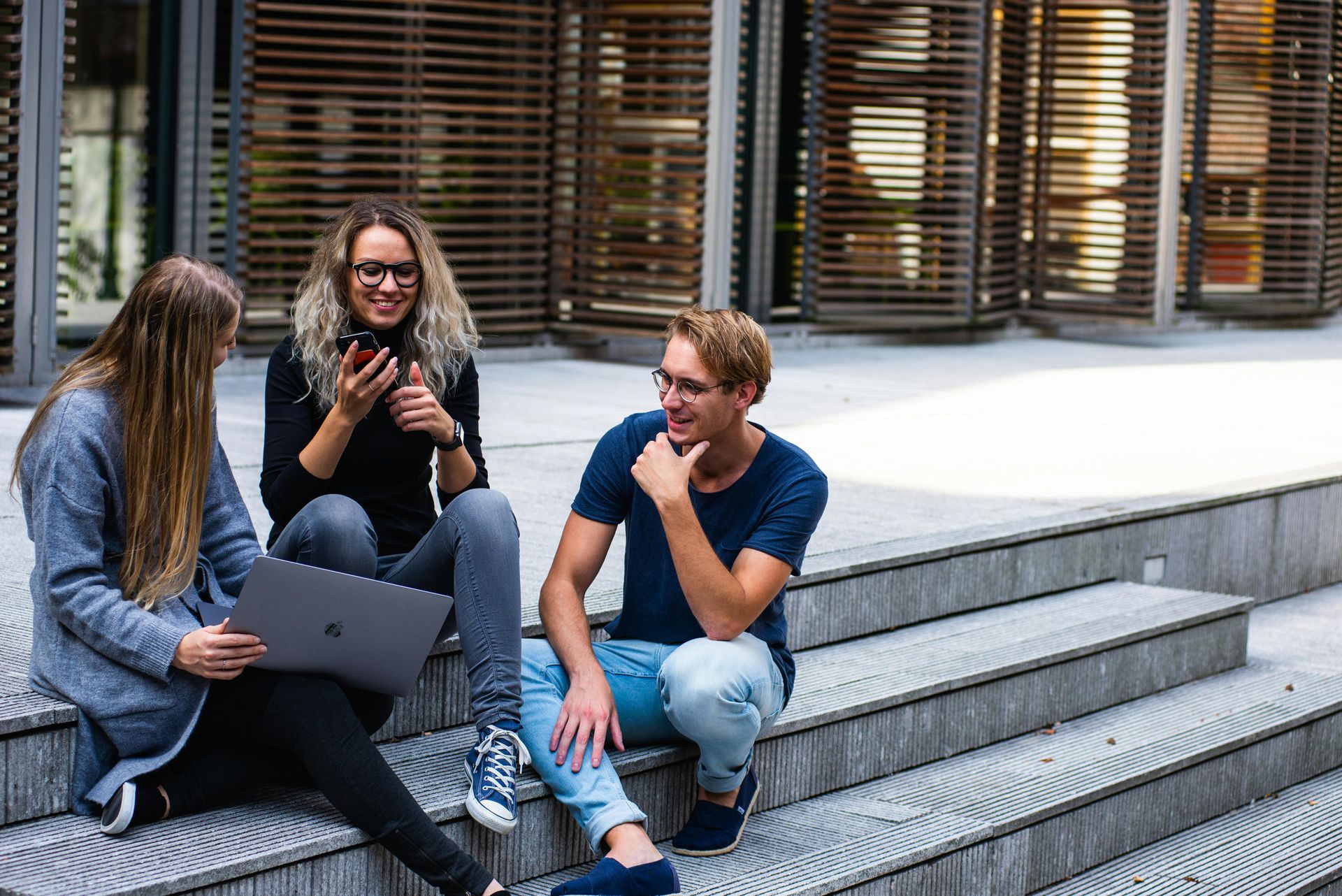 Three young adults sitting on steps outside; one with a laptop, one with a phone, and one smiling, all chatting.