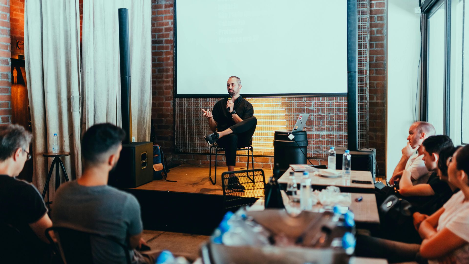 Man giving presentation to audience. He sits on a stool, gesturing. People watch in a room with brick wall.