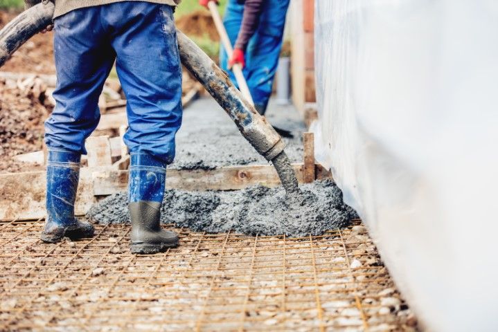 Workers pouring concrete onto a wire mesh foundation outdoors, near a white structure.