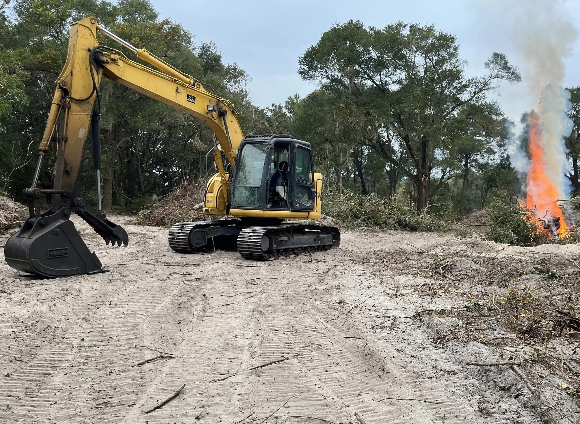 A yellow excavator is driving down a dirt road next to a fire.