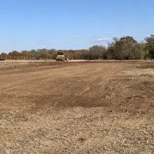 A bulldozer levels a field on a sunny day. Brown dirt and dry grass are visible.