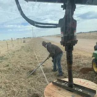 Man using a post driver near a rural road. Brown jacket, jeans, fence line, and overcast sky.