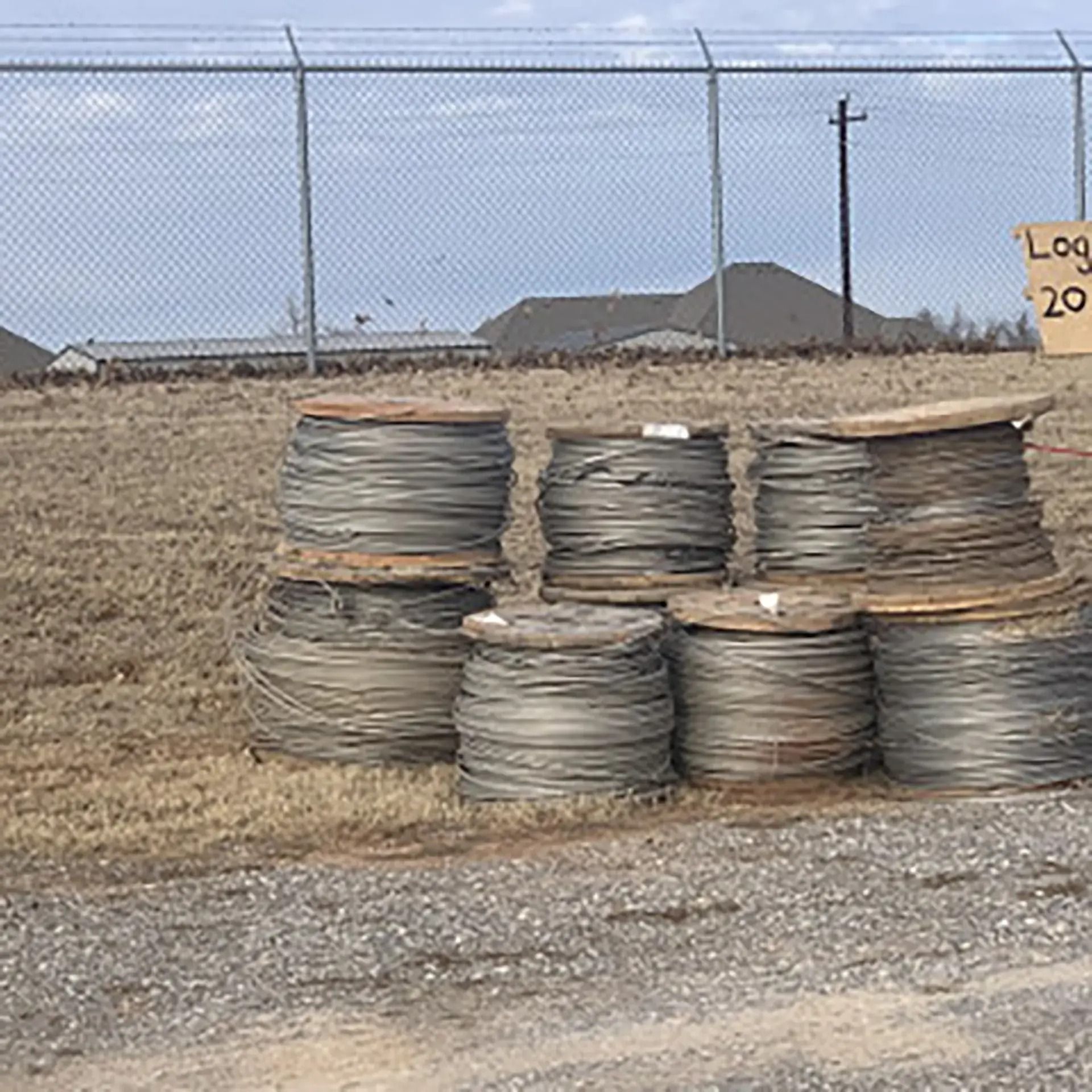 Spools of wire on a grassy field near a fence, houses, and a sign under a cloudy sky.
