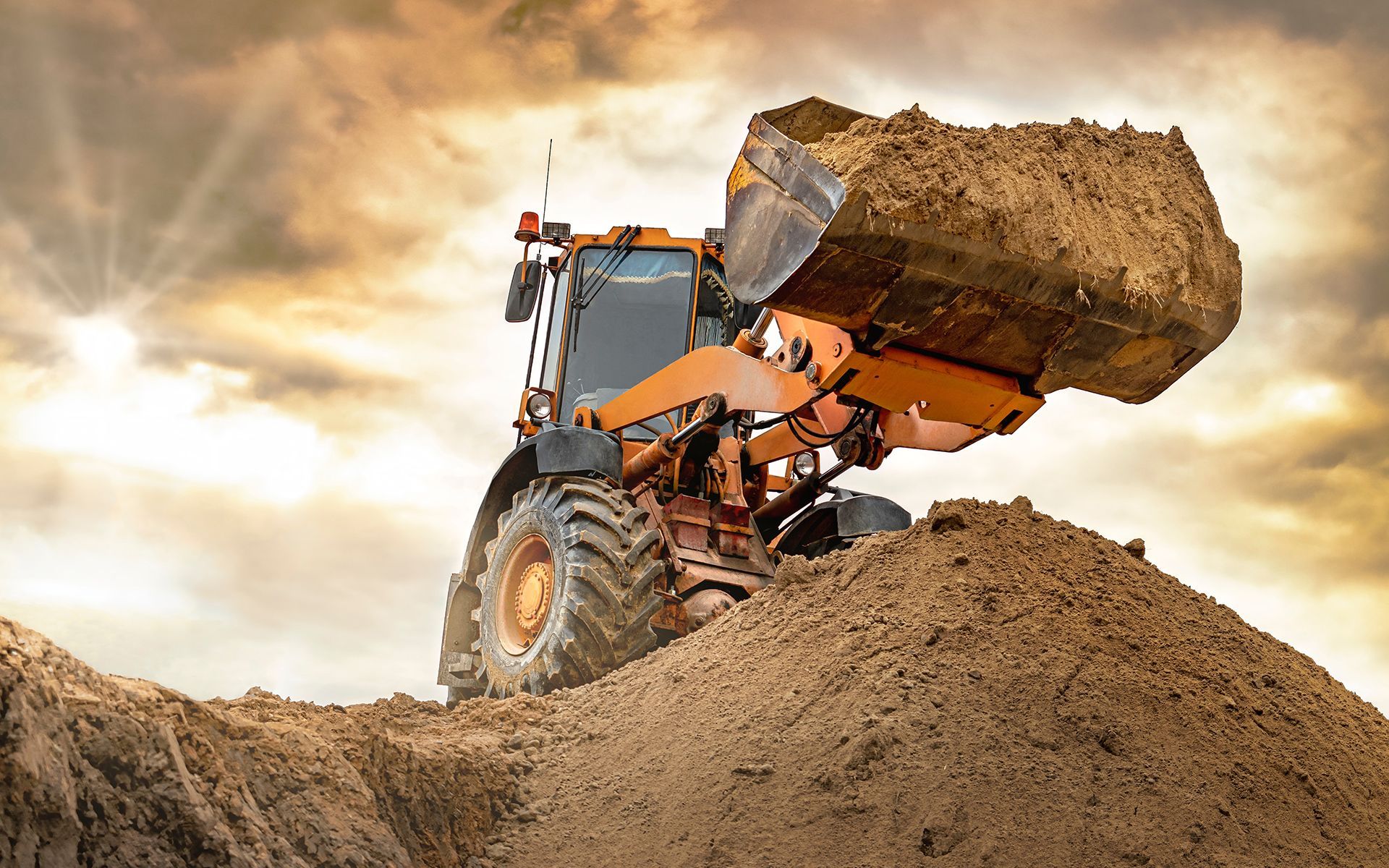 Orange backhoe loader with bucket full of dirt, against a cloudy sky.