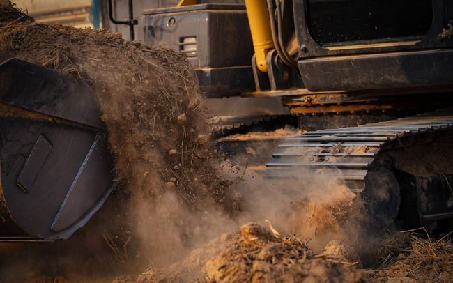 Excavator bucket dumping dirt onto a pile, dusty outdoor setting.