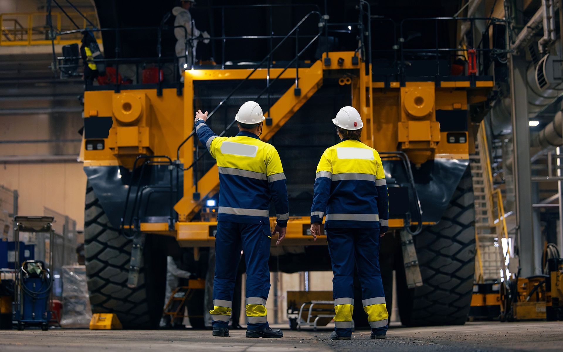 Two workers in safety gear inspect a large yellow mining truck in a factory.