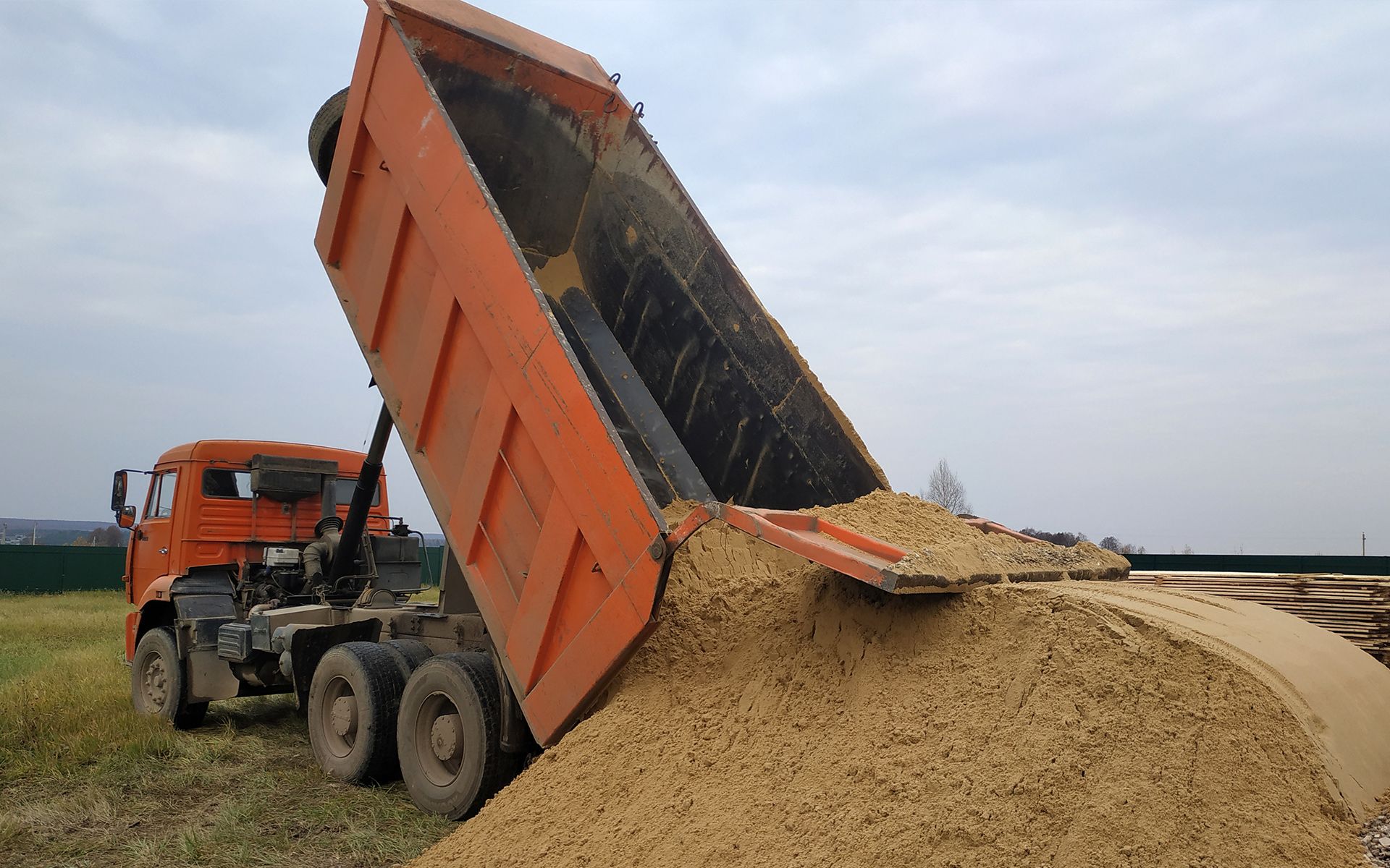 Orange dump truck unloading soil onto a grassy area under a cloudy sky.