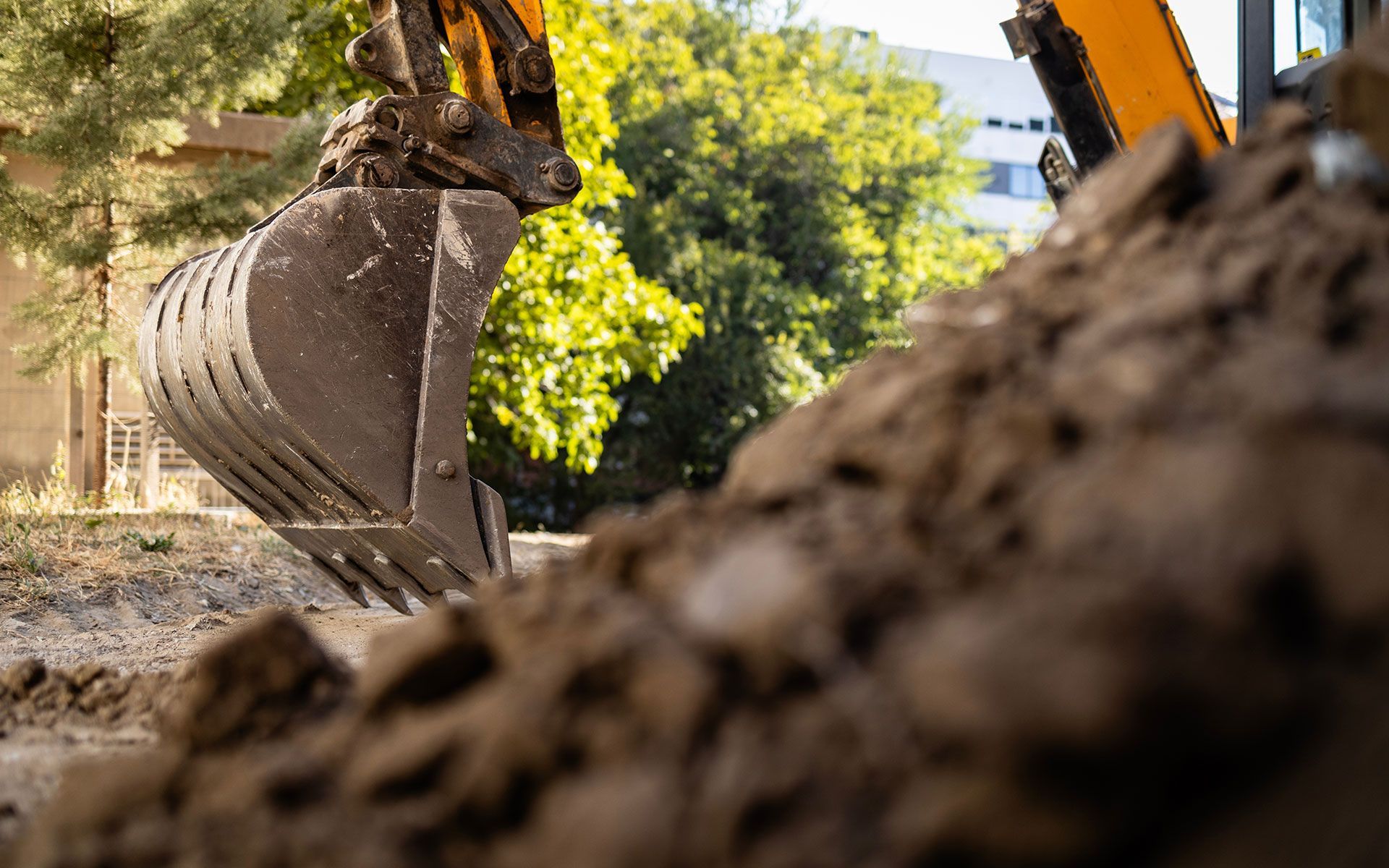 Excavator bucket scoops up dirt at a construction site, trees and building in background.