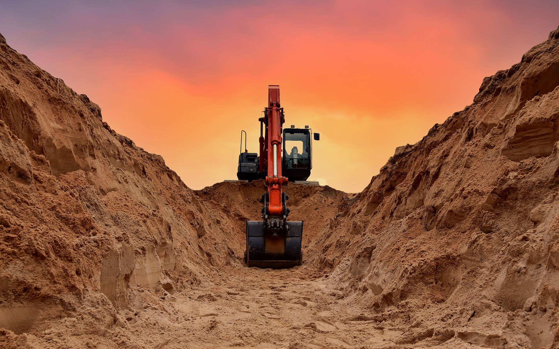 Excavator in a deep trench, digging. Sunset in the background, orange sky.