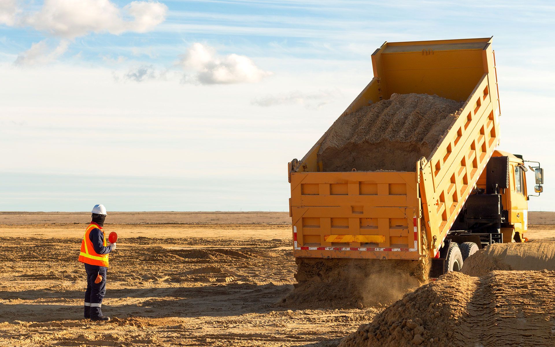 Construction worker directing a yellow dump truck unloading gravel on a barren construction site.