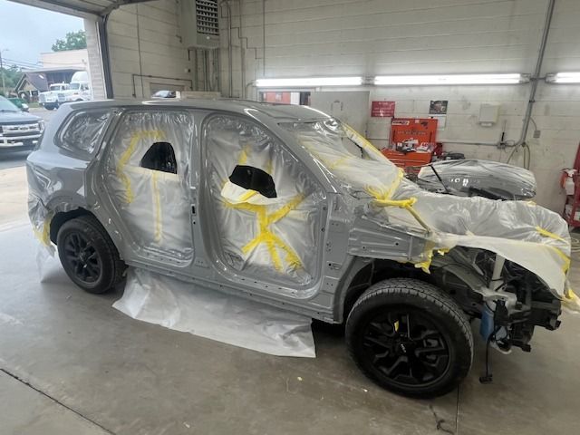 A gray SUV in a body shop, masked for painting. Black wheels, open doors, and exposed front end.
