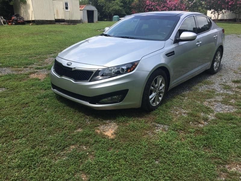 Silver Kia Optima sedan parked on grassy area in front of a house.