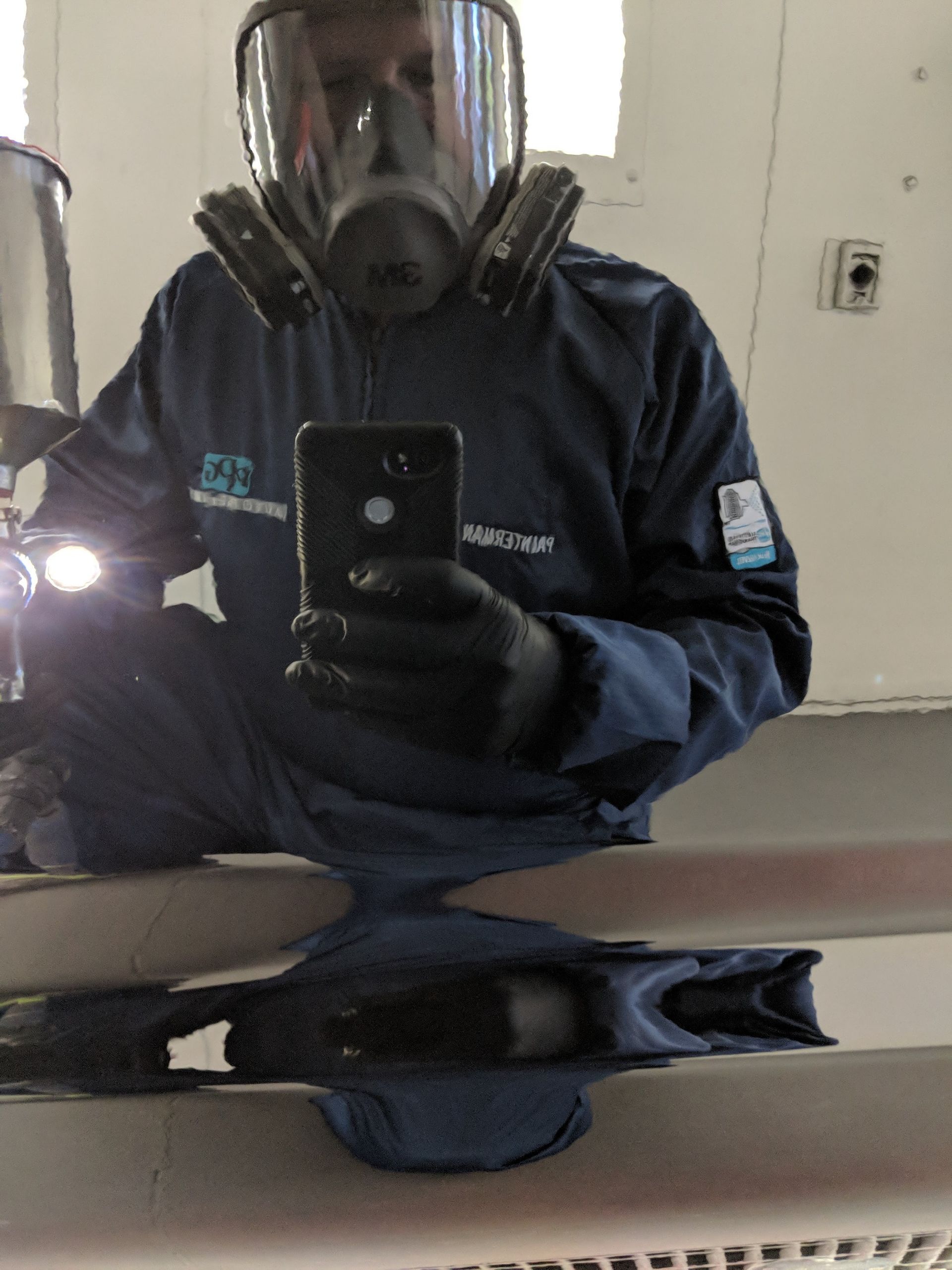 Person in protective gear, holding a phone, reflected in a shiny surface; inside a white industrial setting.