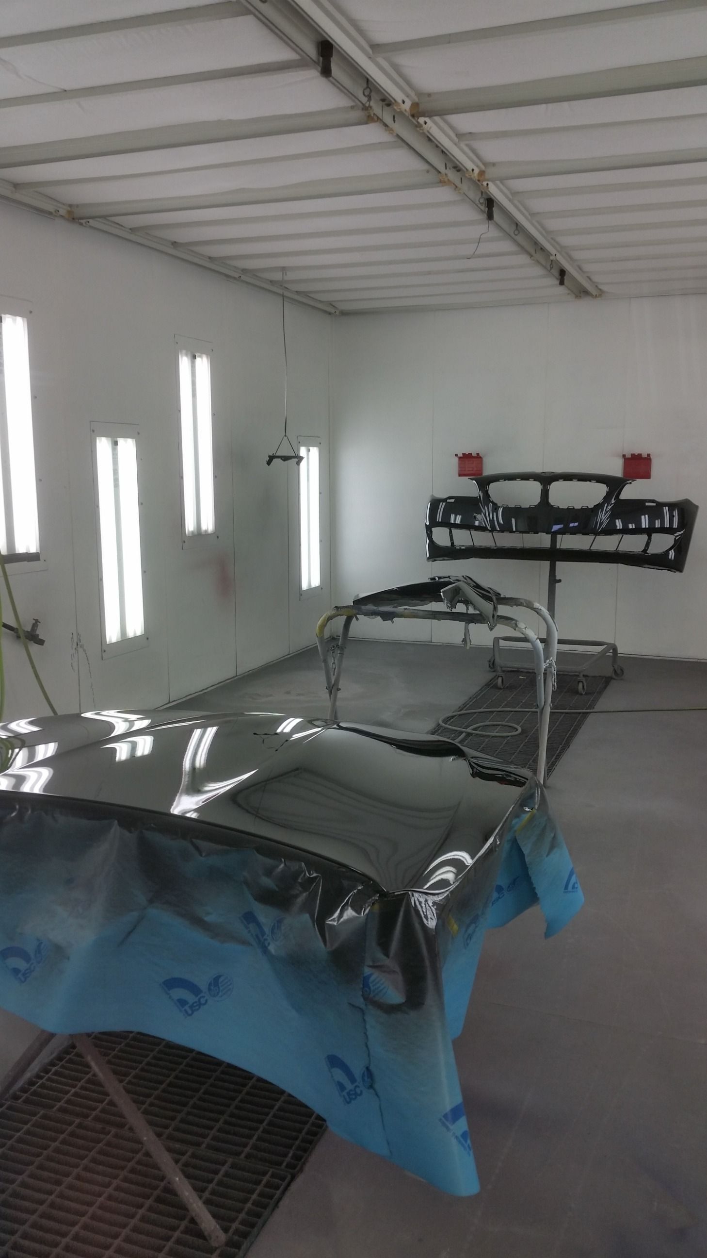 Inside a paint booth; car parts covered and on stands; bright lights illuminate the room.