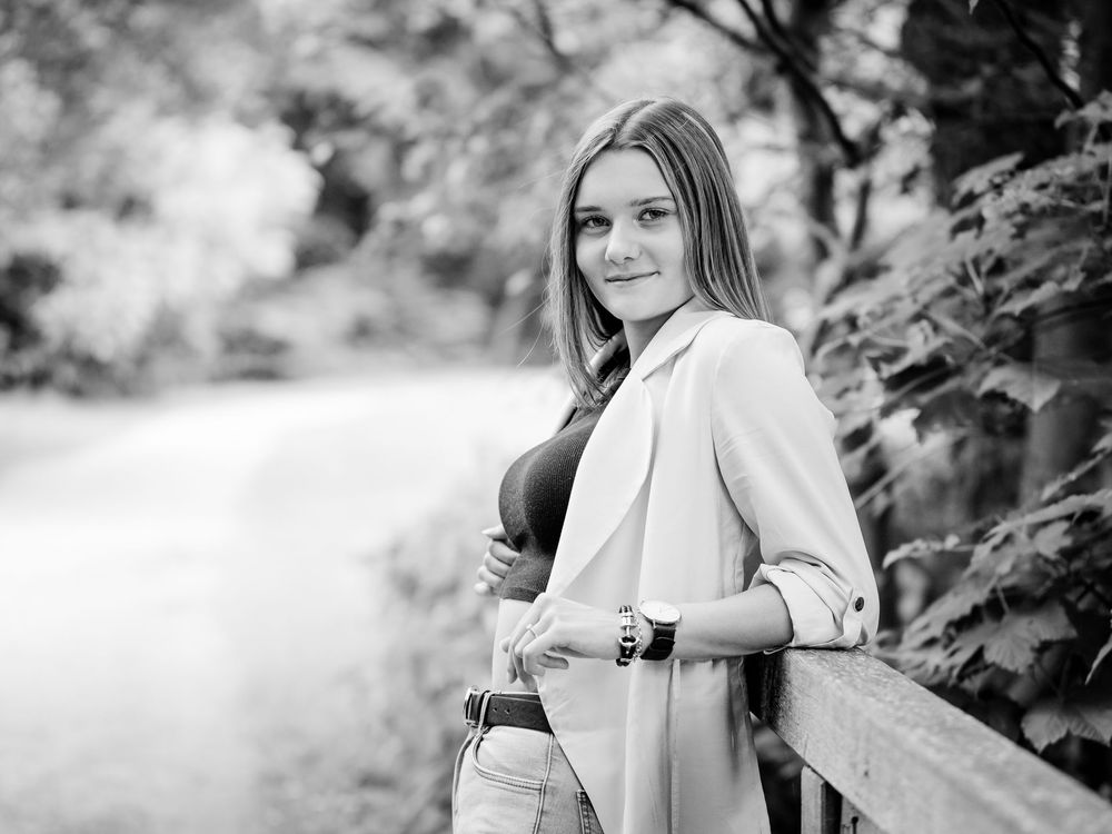 Junge Frau lehnt im Freien an einem Holzzaun und lächelt auf einem Schwarz-Weiß-Porträt.