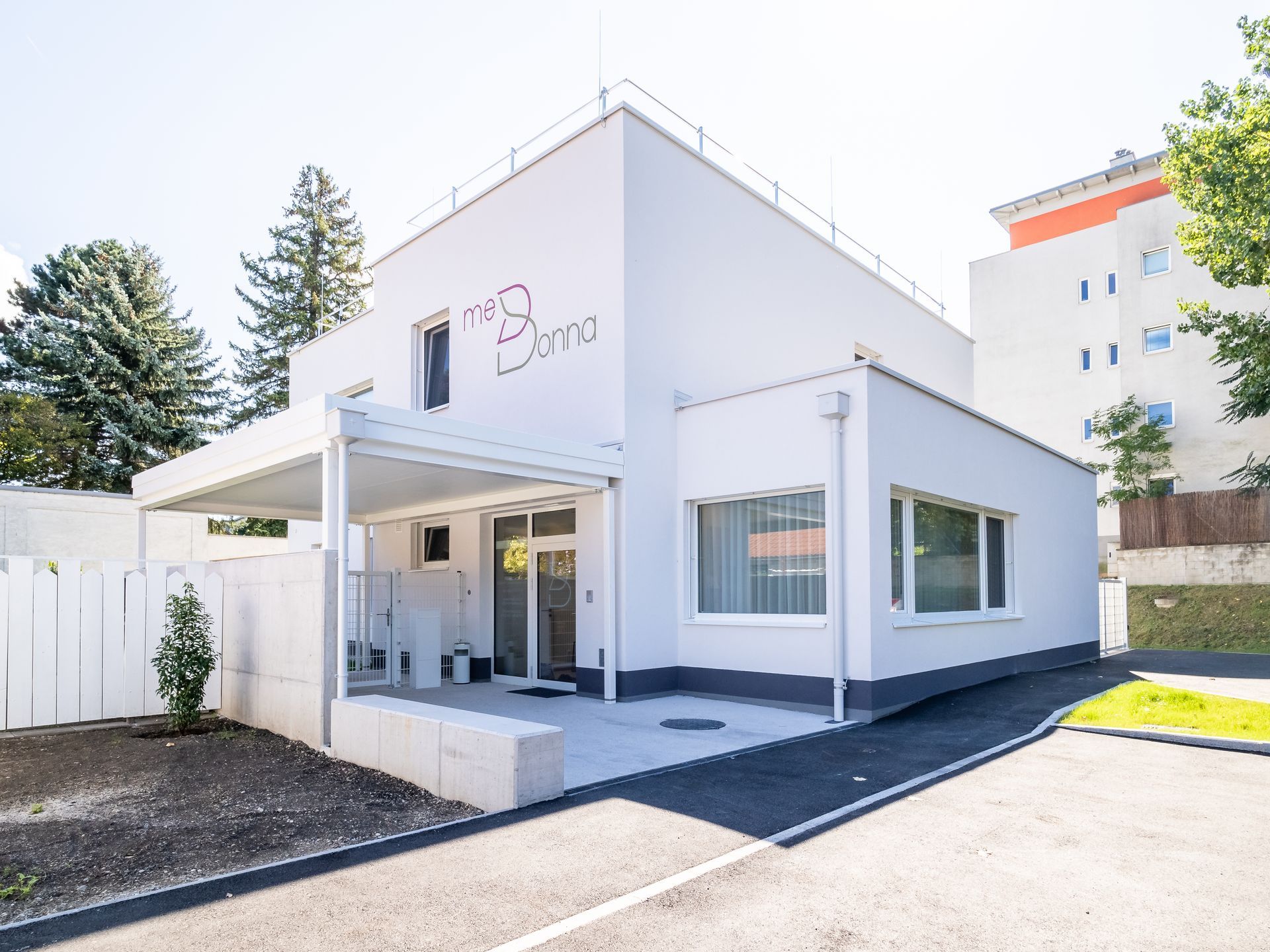 Modernes, weißes Gebäude mit rosa Logo, Haupteingang und Auffahrt in einem sonnigen Wohngebiet