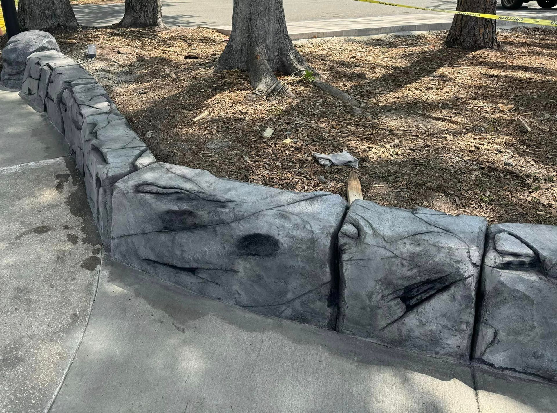 A stone wall along a sidewalk in a park with trees in the background.