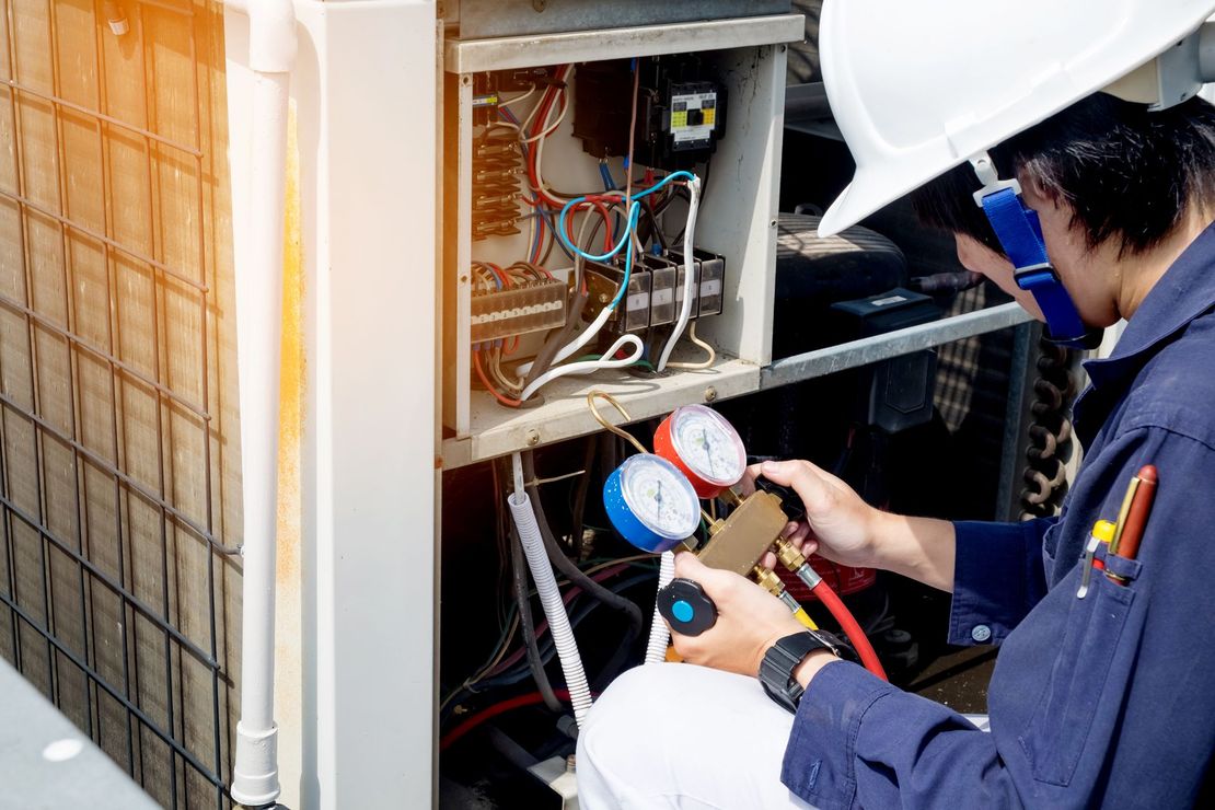 HVAC technician in a white hard hat and blue jumpsuit, working on an outdoor air conditioning unit, using gauges.