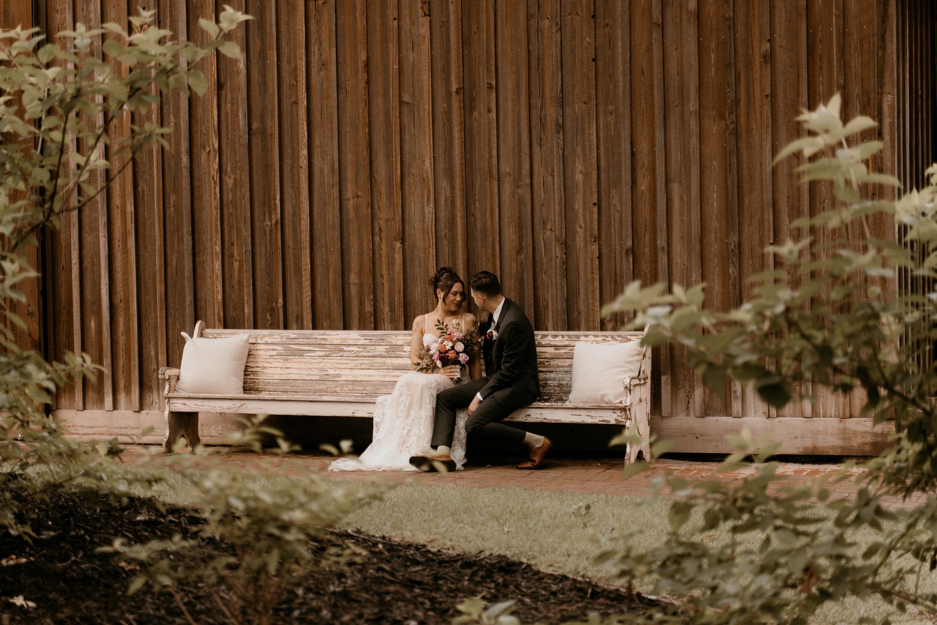 A bride and groom are sitting on a bench in front of a wooden wall.